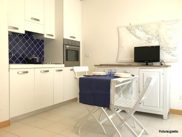 A compact kitchen with white cabinets, a stove, and a microwave. There's a dining table with a blue tablecloth and a chair. A television is mounted on the wall above the table. - Cozy Nest for Your Getaway in Grado (Grado)