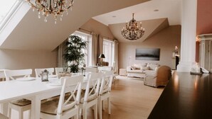 Ein geräumiger Essbereich mit hoher Decke und einer Chandelier, ein gemütlicher Wohnbereich mit Sofa und Fernseher sowie ein großes Fenster, das für natürliches Licht sorgt.