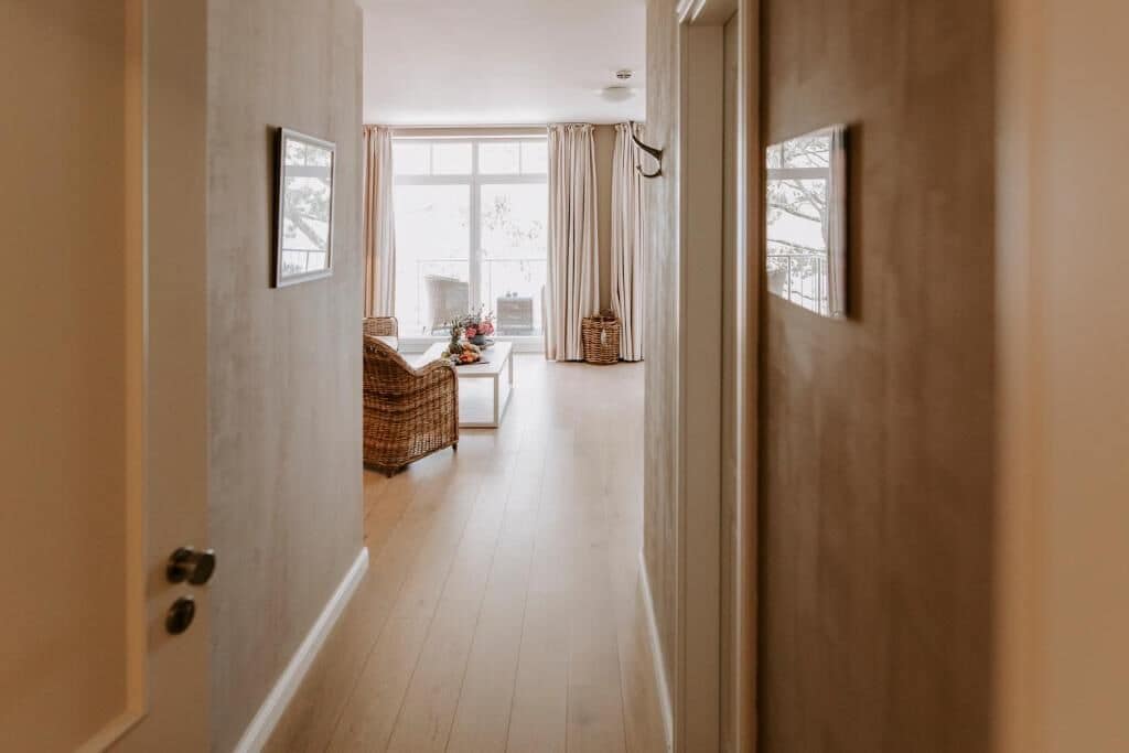 Ein Flur, der zu einem Zimmer mit Korbsessel, einem kleinen Tisch mit Blumen und einem fenster mit Vorhängen führt.