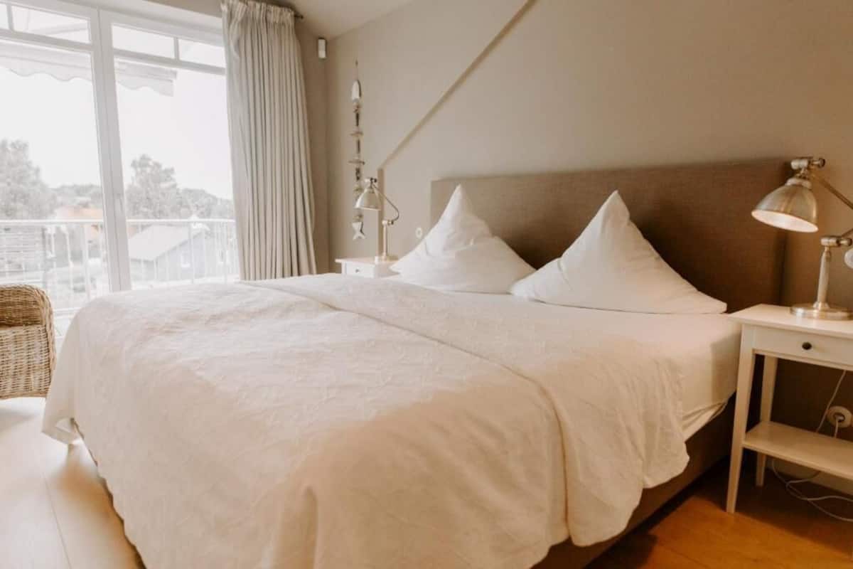 Ein Schlafzimmer mit einem großen Bett, zwei Nachttischen, einem Fenster mit Vorhängen und Blick ins Freie.