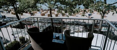 A balcony with wicker chairs and a table, overlooking a beach with sun loungers and bicycles.