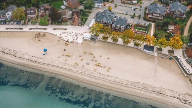 Luftaufnahme einer Küstenstadt mit Sandstrand, einer Reihe von Häusern und einem markanten Gebäude mit dunklem Dach.