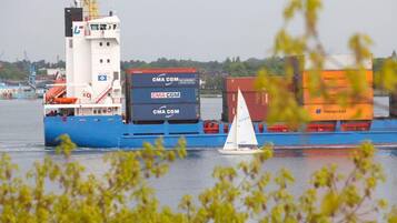 Un grand cargo transportant de nombreux conteneurs, un petit voilier et un navire amarré en arrière-plan.