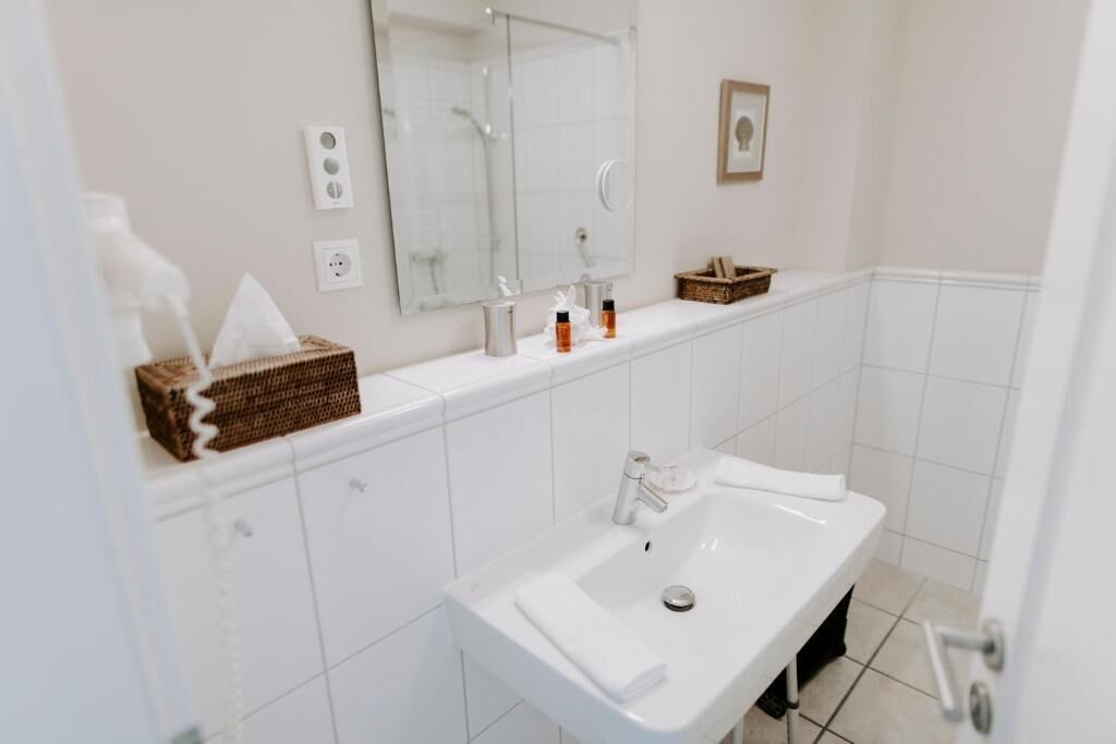 A modern bathroom with a white sink, a mirror, and a shower. There are toiletries on the counter and a basket of towels.