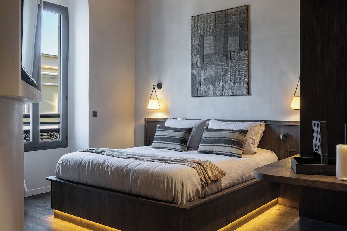 A modern bedroom with a large bed, two bedside lamps, a wall-mounted artwork, and a window with blinds.