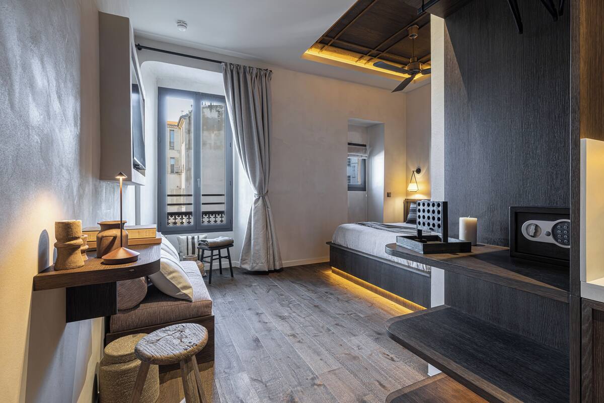 A modern hotel room with a wooden floor, a bed with a headboard, a desk with a lamp, a chair, and a window with curtains.