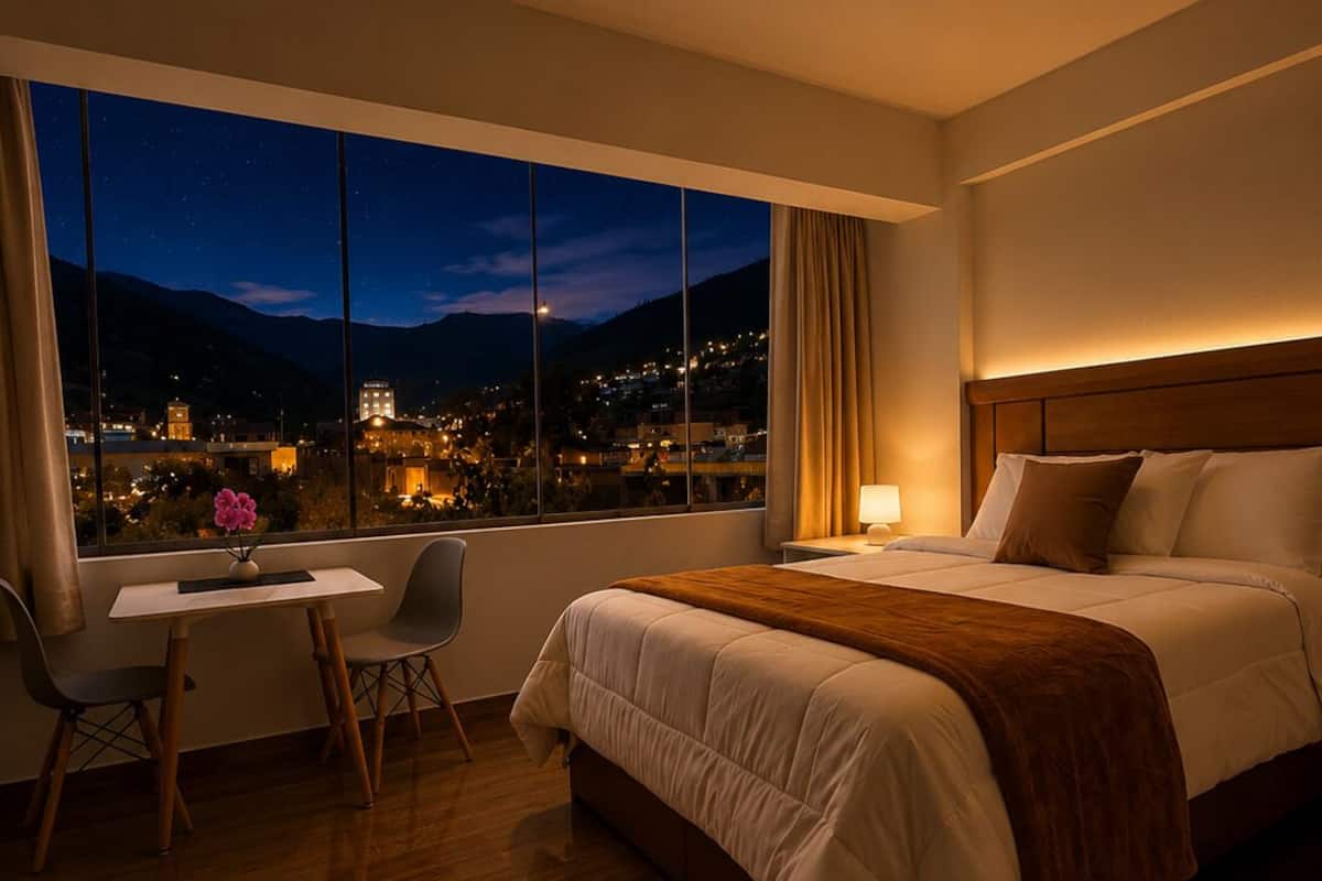 A hotel room with a large window, a bed, a desk with a chair, and a nightstand.