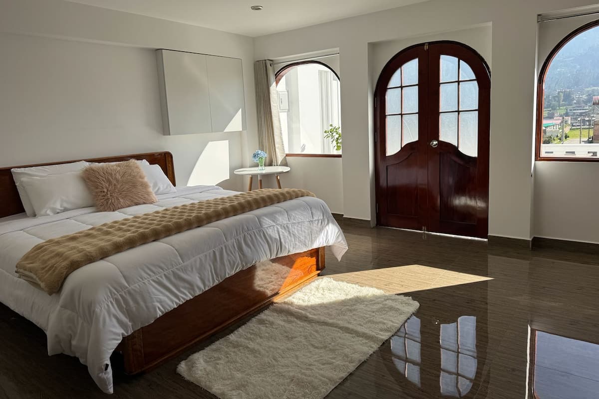 A bedroom with a large bed, a wooden headboard, a white rug, and a view of the city through a large arched window.
