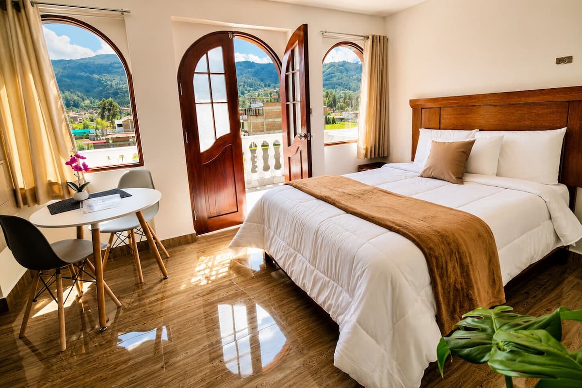 A hotel room with a bed, a desk, and chairs, large windows with a view of greenery and mountains.