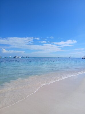 Ein Strand mit klarem, blauem Wasser, einem sandigen Ufer und mehreren Booten in der Ferne.
