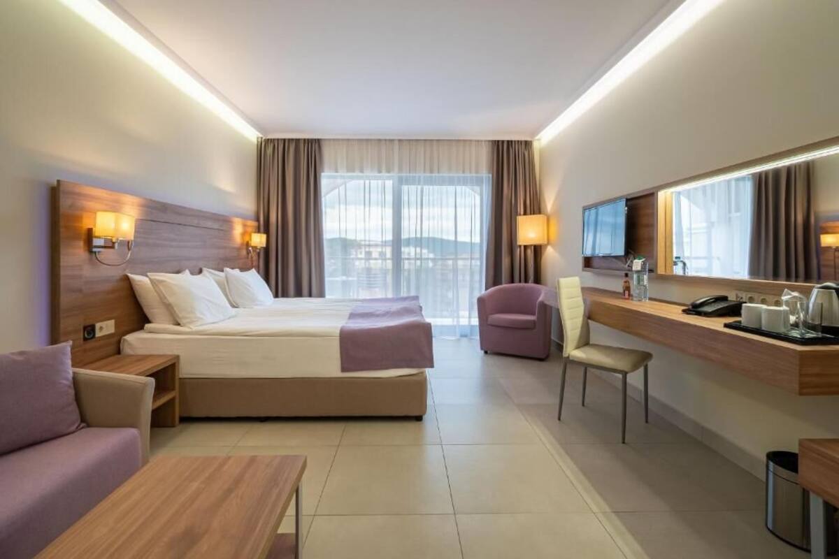A modern hotel room with a large bed, a desk with a chair, a sofa, and a TV mounted on the wall.