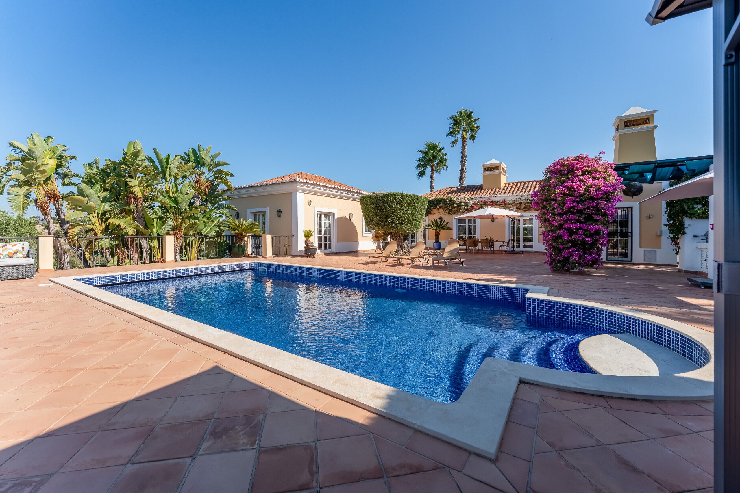 A swimming pool with tiled edges, surrounded by palm trees and a patio area with outdoor furniture.