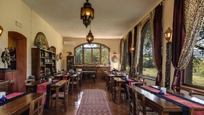 Uma área de refeições com mesas e cadeiras de madeira, uma janela grande com vista para o verde e uma luminária decorativa no teto.