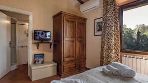 Quarto com cama, guarda-roupa de madeira, televisão em uma estante, um livro e janela com vista para árvores.