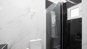 Un baño moderno con ducha envuelta en vidrio, paredes de mármol y un inodoro con dispensador de toallas de papel.