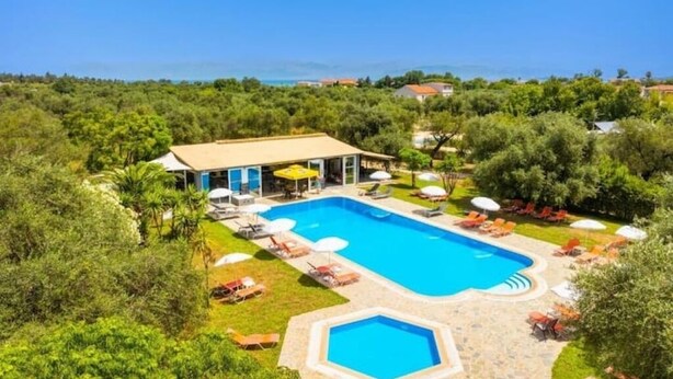 Vista aérea de una propiedad con una amplia piscina rectangular rodeada de sillones de descanso y sombrillas, frente a un telón de fondo de vegetación exuberante y un cielo azul despejado.