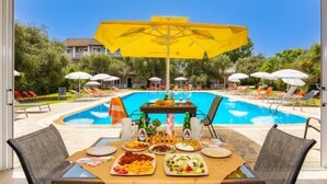 Una mesa de comedor con comida y bebidas, una piscina con sillones de descanso y sombrillas en un entorno de resort.