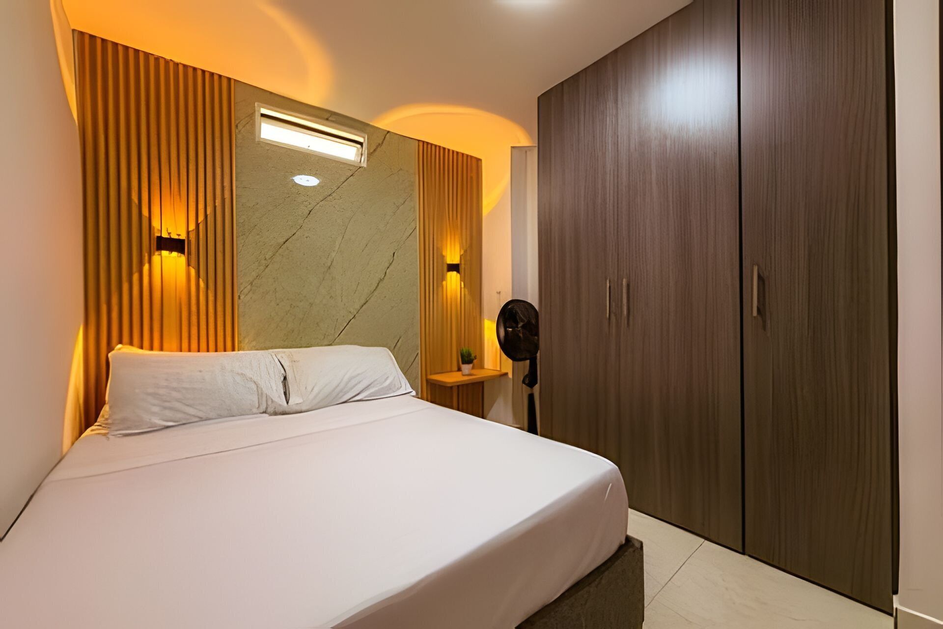 A hotel room with a bed, a wardrobe, a nightstand, and a wall-mounted fan.