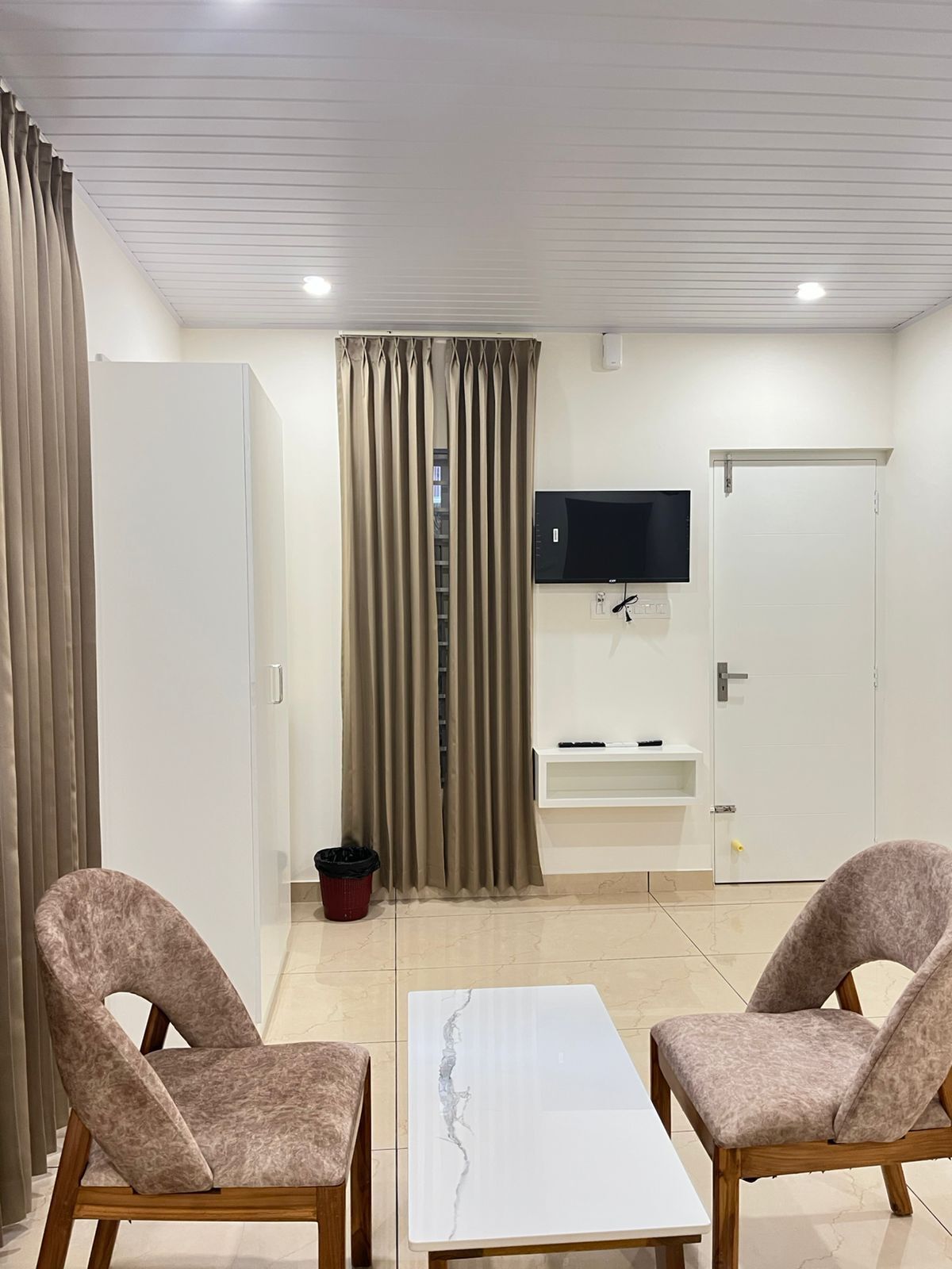 A modern living room with two armchairs, a white coffee table, a flat-screen TV mounted on the wall, a sliding door, and a small shelf with a remote control.
