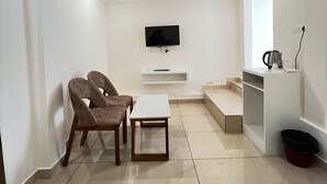 A modern living room with a flat-screen TV mounted on the wall, a sofa, a coffee table, and a small kitchen area with a kettle and a trash bin. - aagneyam grand RESORT (Kozhikode)