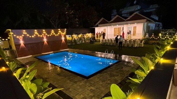A swimming pool surrounded by plants and string lights, with a building in the background. - aagneyam grand RESORT (Kozhikode)