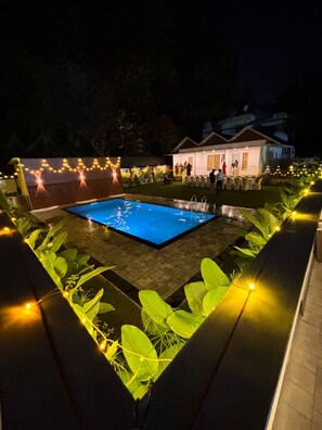 A swimming pool surrounded by plants and string lights, with a building in the background. - aagneyam grand RESORT (Kozhikode)