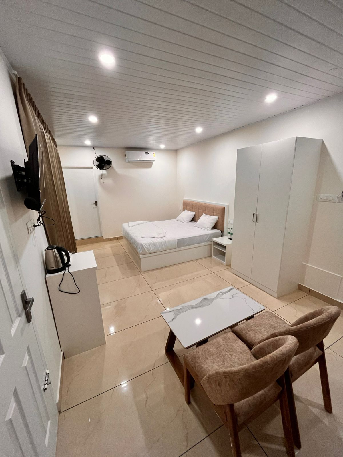 A modern bedroom with a bed, bedside table, wardrobe, and a small dining area with chairs.