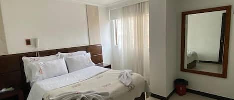 Een hotelkamer met een bed, nachtkastje, spiegel en een raam met gordijnen.