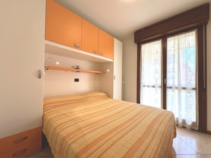 Ein Schlafzimmer mit einem Bett, einem orangefarbenen Kleiderschrank, einem Regal über dem Bett und einem Fenster mit Vorhängen.