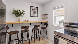 Une cuisine avec un comptoir-bar en bois, trois tabourets, une plante en pot, un tableau encadré et un four en acier inoxydable.