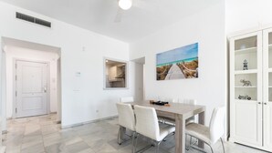 A dining area with a wooden table and white chairs, a white cabinet with glass doors, and a beach-themed painting on the wall. - Apartment '203 Arruzafa Sea View' with Sea View, Wi-Fi and Air Conditioning (Las Lagunas de Mijas)