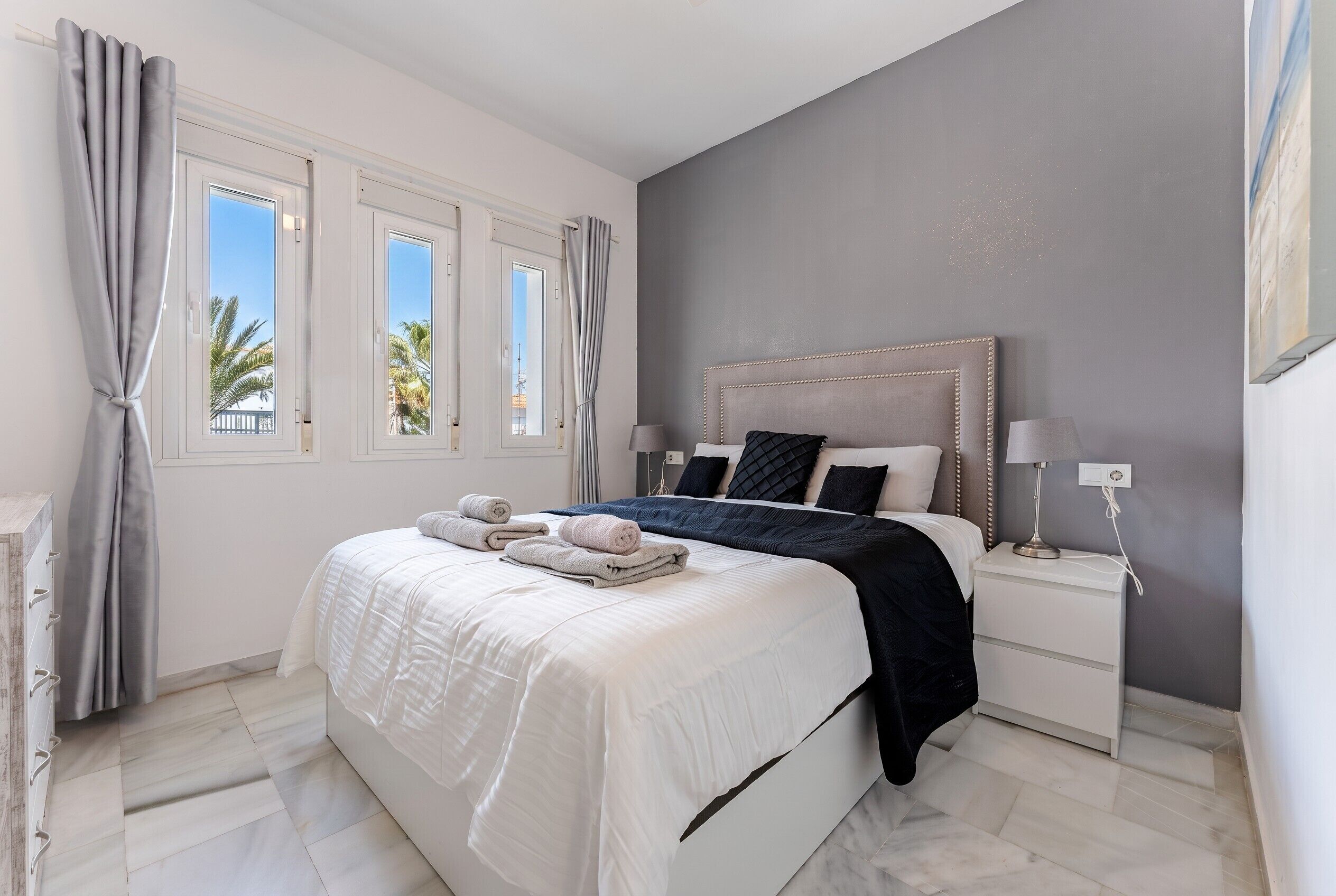 Un dormitorio con cama, mesitas de noche, lámpara y vistas a palmeras a través de la ventana.