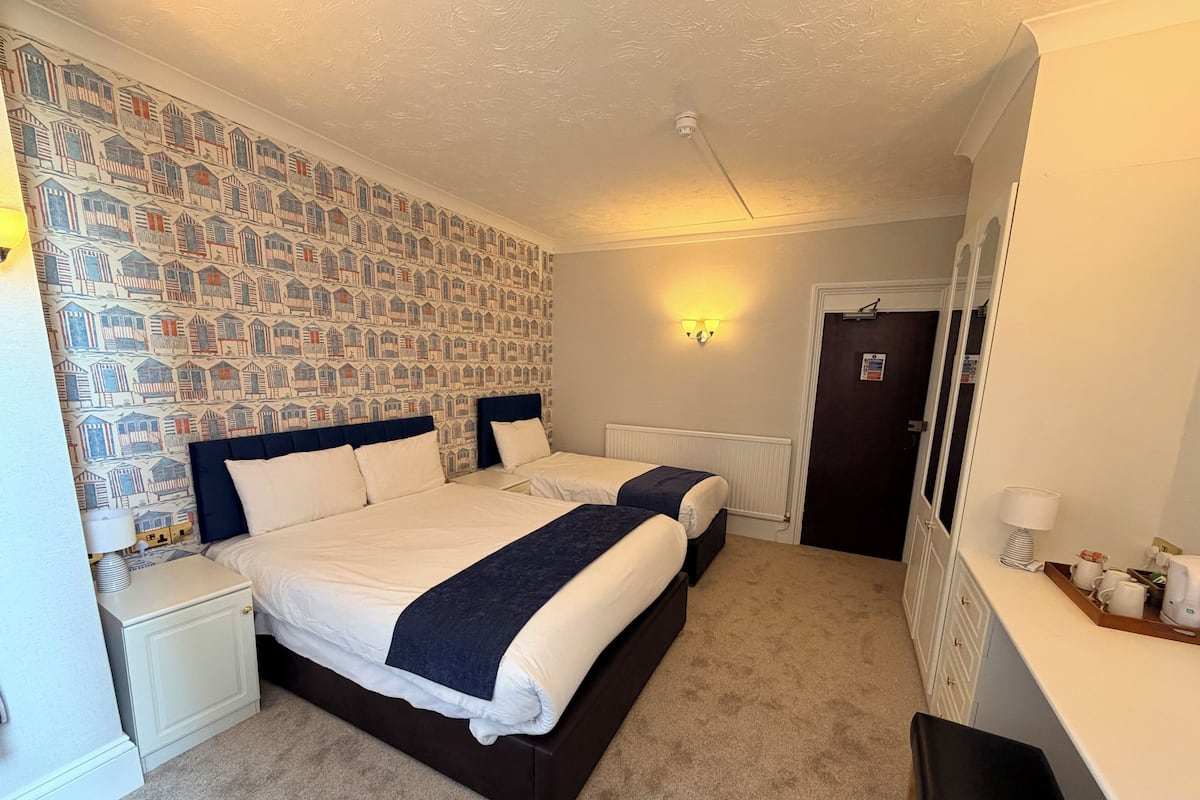 A hotel room with two beds, a patterned wallpaper, a radiator, a desk with a lamp, and a door with a sign.