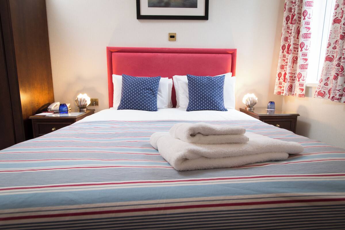 A hotel room with a bed, pillows, a nightstand, a lamp, and a window with curtains.
