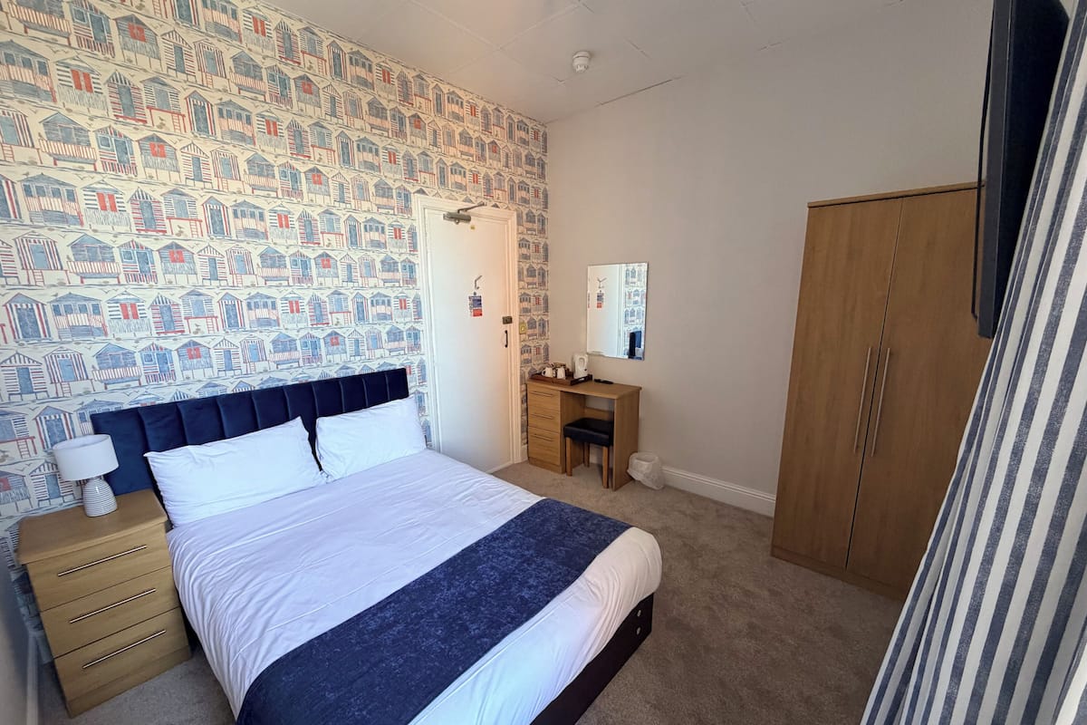 A bedroom with a patterned wallpaper, a bed with white and dark blue bedding, a wooden dresser, a desk, and a wardrobe.
