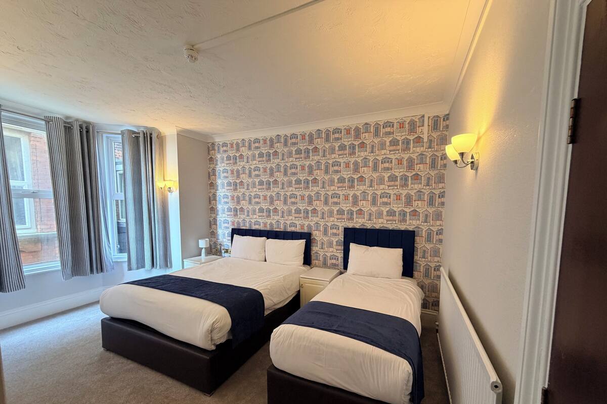 A double room with two beds, a patterned wallpaper, a window with curtains, and a wall-mounted light.