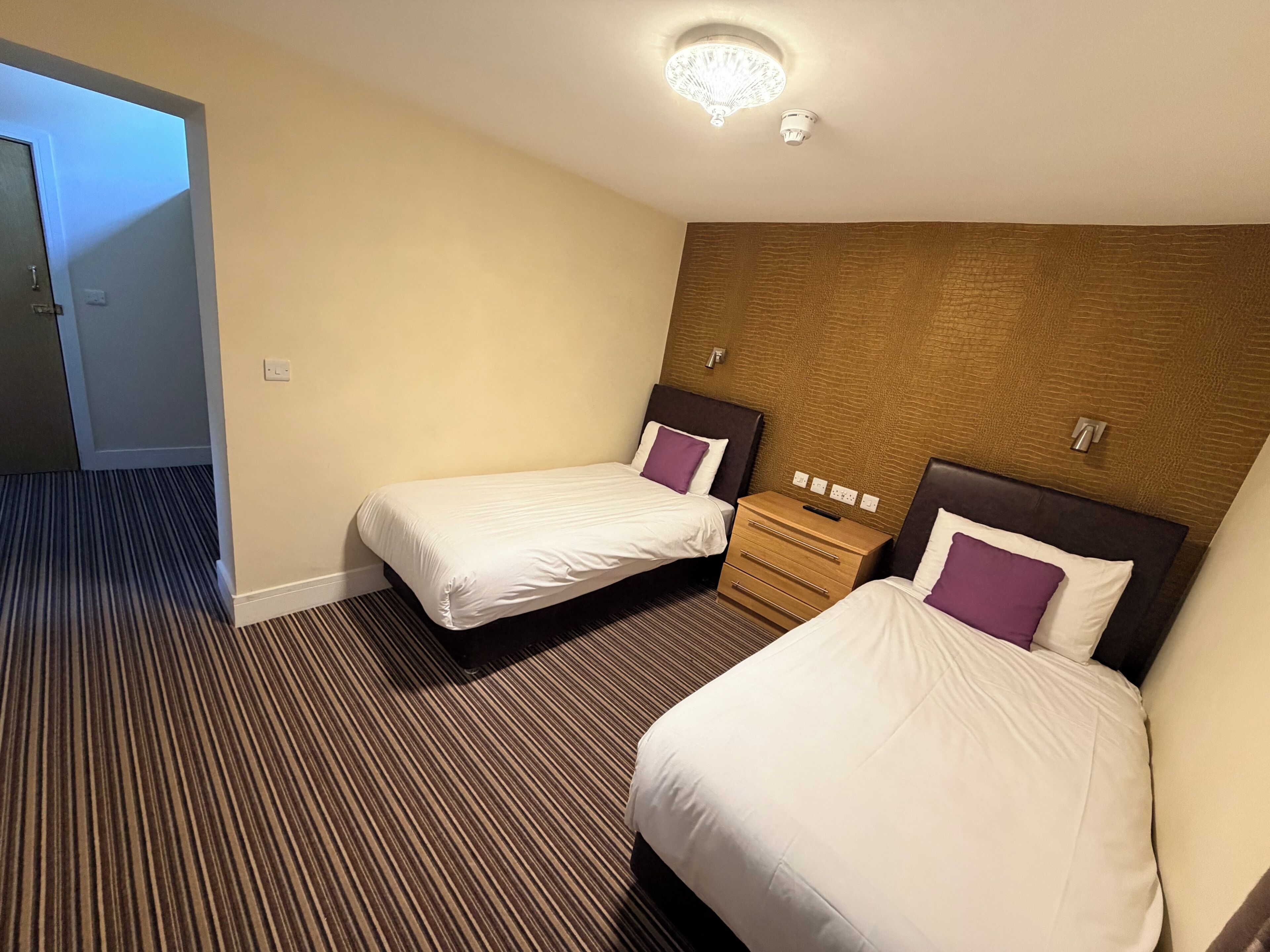 Une chambre d’hôtel avec deux lits, une tête de lit en bois, un sol recouvert de moquette et un luminaire au plafond.