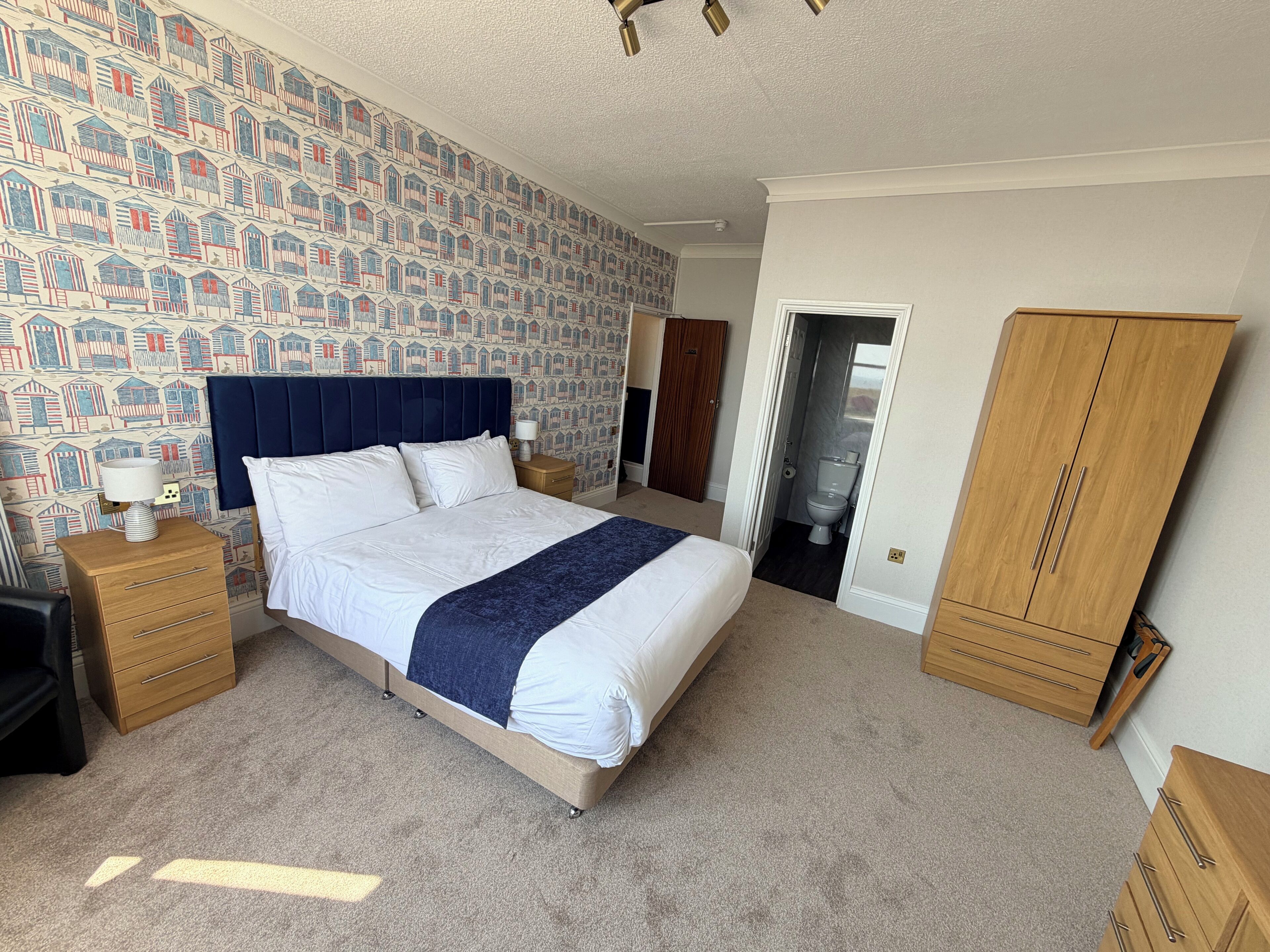 A bedroom with a bed, wooden nightstands, a wardrobe, and a patterned wallpaper.