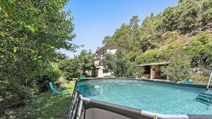 A swimming pool with a wooden deck, surrounded by greenery and a house in the background. - Holiday Home 'Maison En Cevennes' with Mountain View, Private Pool and Private Terrace (L'Estréchure)