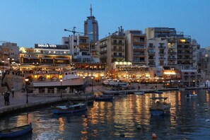 Une vue sur la ville, avec ses bâtiments qui bordent le front de mer, dont un bâtiment particulièrement imposant portant l’inscription « DOLCE VITA », et un port animé où de nombreux bateaux sont amarrés.