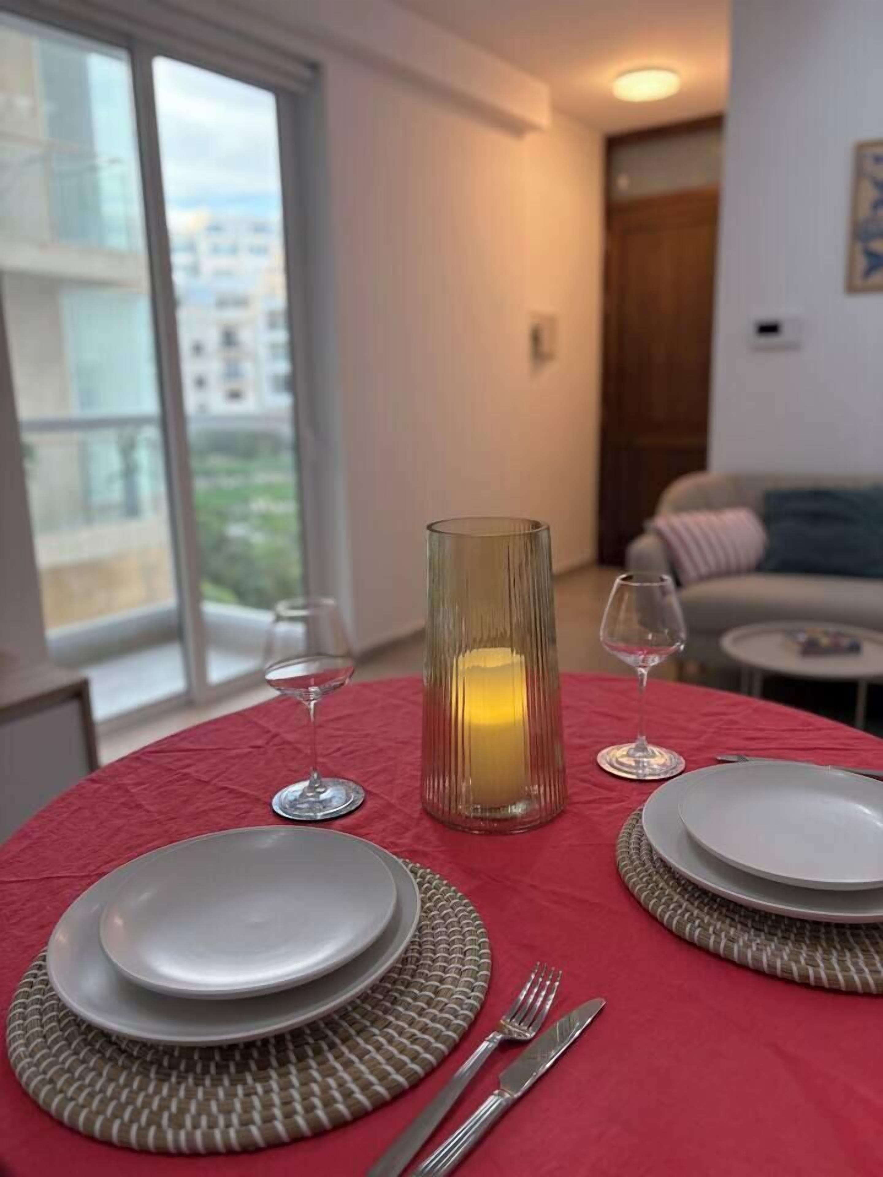A dining area with a table set for two, featuring a candle centerpiece, a red tablecloth, and a view of the city through large windows.