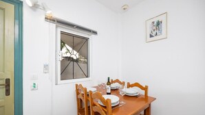 Un coin repas avec une table en bois dressée pour deux personnes, une fenêtre ornée d’une grille métallique et un tableau encadré accroché au mur.