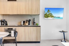 A modern kitchen with wooden cabinets, a Bosch oven, a coffee machine, and a small dining table with a chair. A flat-screen TV mounted on the wall displays a tropical beach scene. - 19th Floor | Luxury 1BR + Den | Rooftop Pool & Gym (Miami)