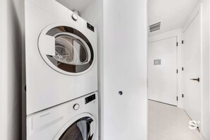 Uma área de lavanderia com duas máquinas de lavar frontais empilhadas e uma porta que dá para outro cômodo.