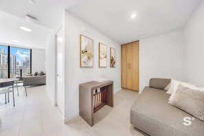 Uma sala de estar moderna com um sofá cinzento, uma mesa pequena e um armário de madeira. Há três quadros abstratos na parede, e uma janela grande oferece vista da cidade.