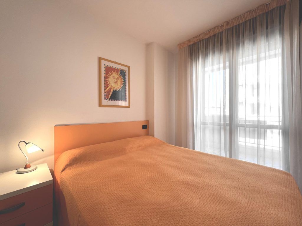A hotel room with a single bed, a nightstand with a lamp, a framed picture on the wall, and a large window with sheer curtains.