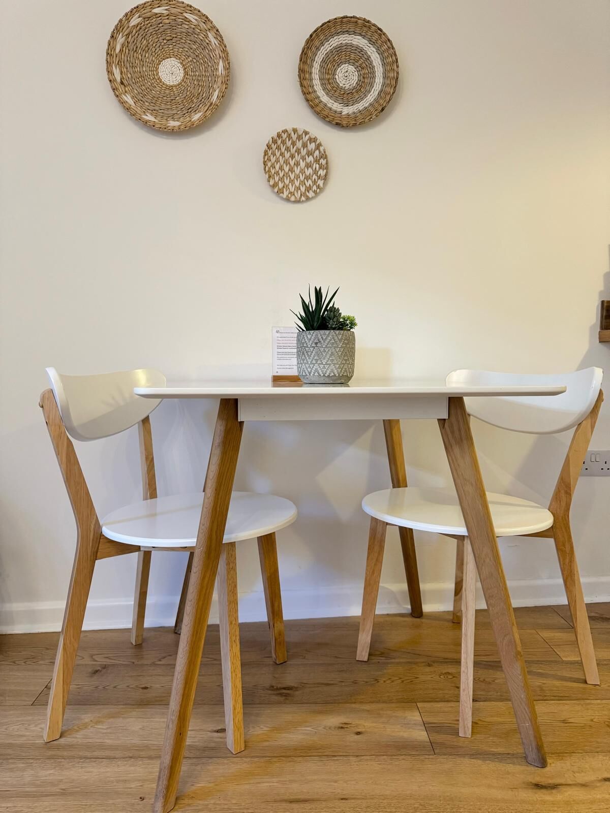 Een eettafel met twee witte stoelen, een kleine potplant en drie geweven wanddecoraties.