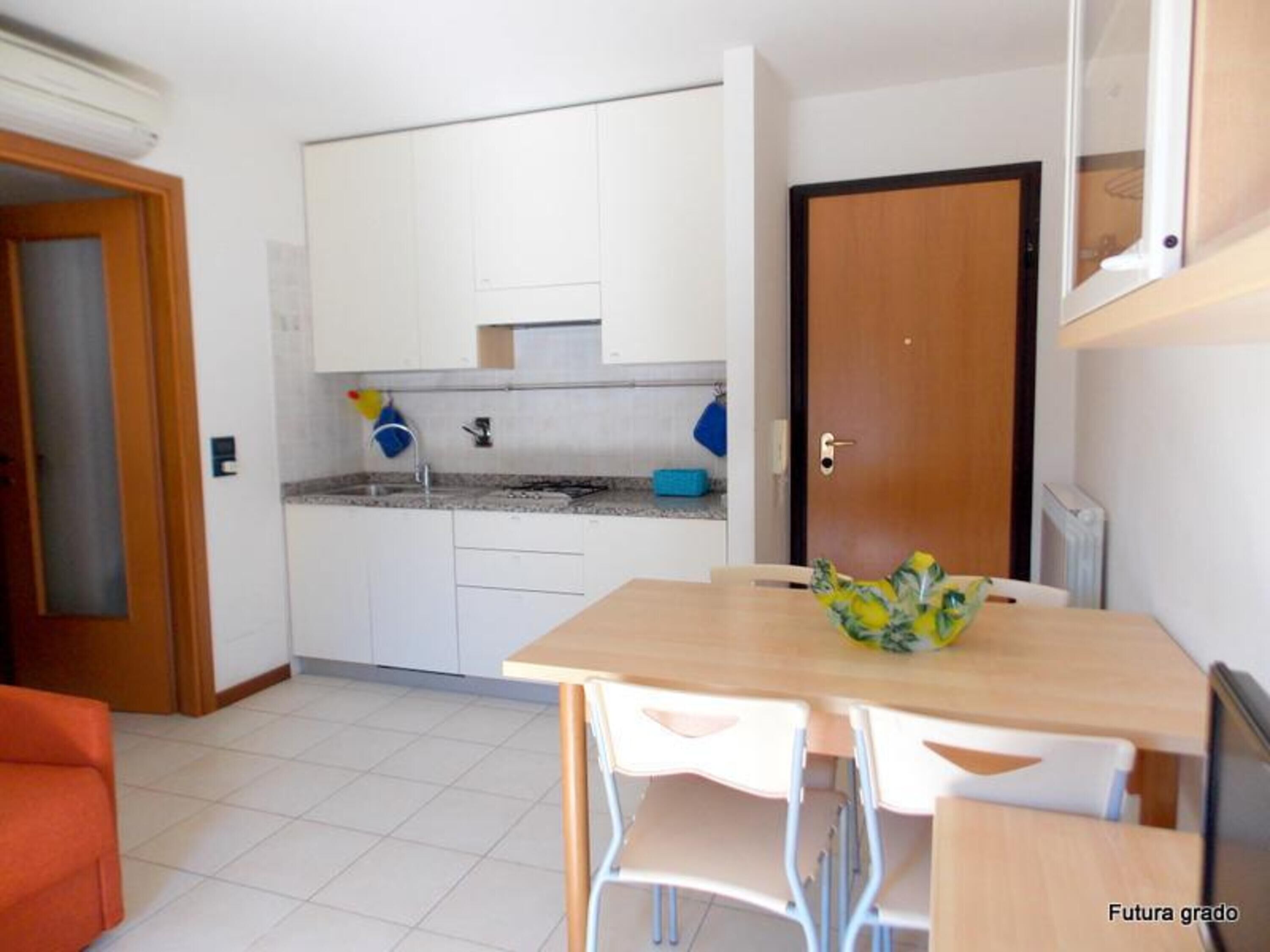 Cocina pequeña con armarios blancos, un lavabo y una encimera. Se aprecia una mesa de comedor de madera con sillas y un cuenco de fruta.