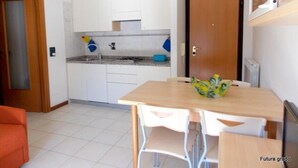 A compact kitchen with white cabinets, a sink, and a countertop. There is a wooden dining table with chairs and a bowl of fruit. - Inviting Apartment Close to the Beach in Grado (Grado)