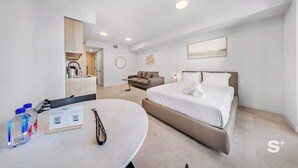 Una habitación de hotel moderna con cama, sofá, mesa de comedor con botellas de agua y una cocina al fondo.